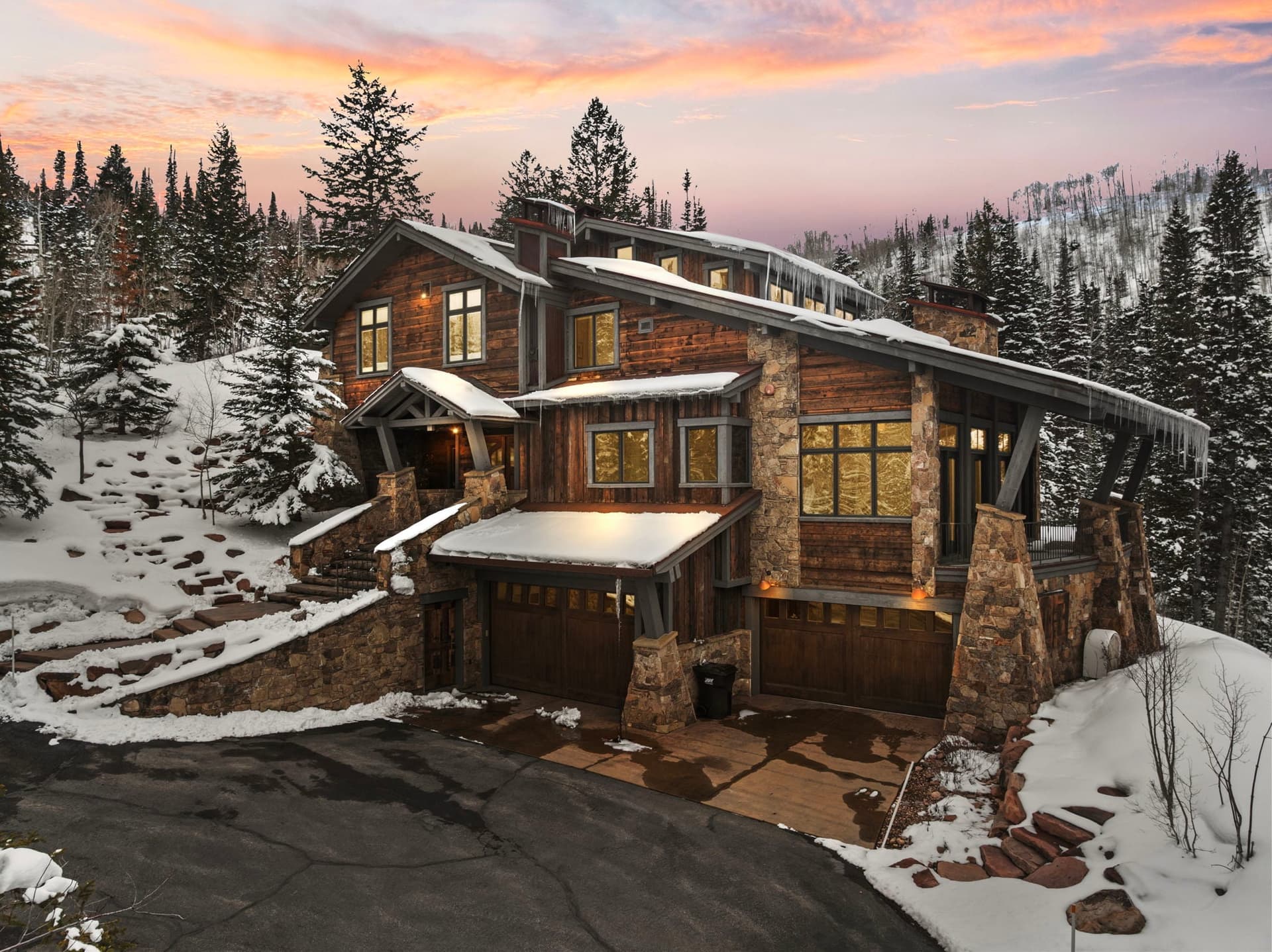 A view of Silver Horse at The Colony in Park City. The home is sitting next to a ski slope, and the windows are glowing with light. Snow covered trees surround it and there is a view of the mountain and sunset in in the background